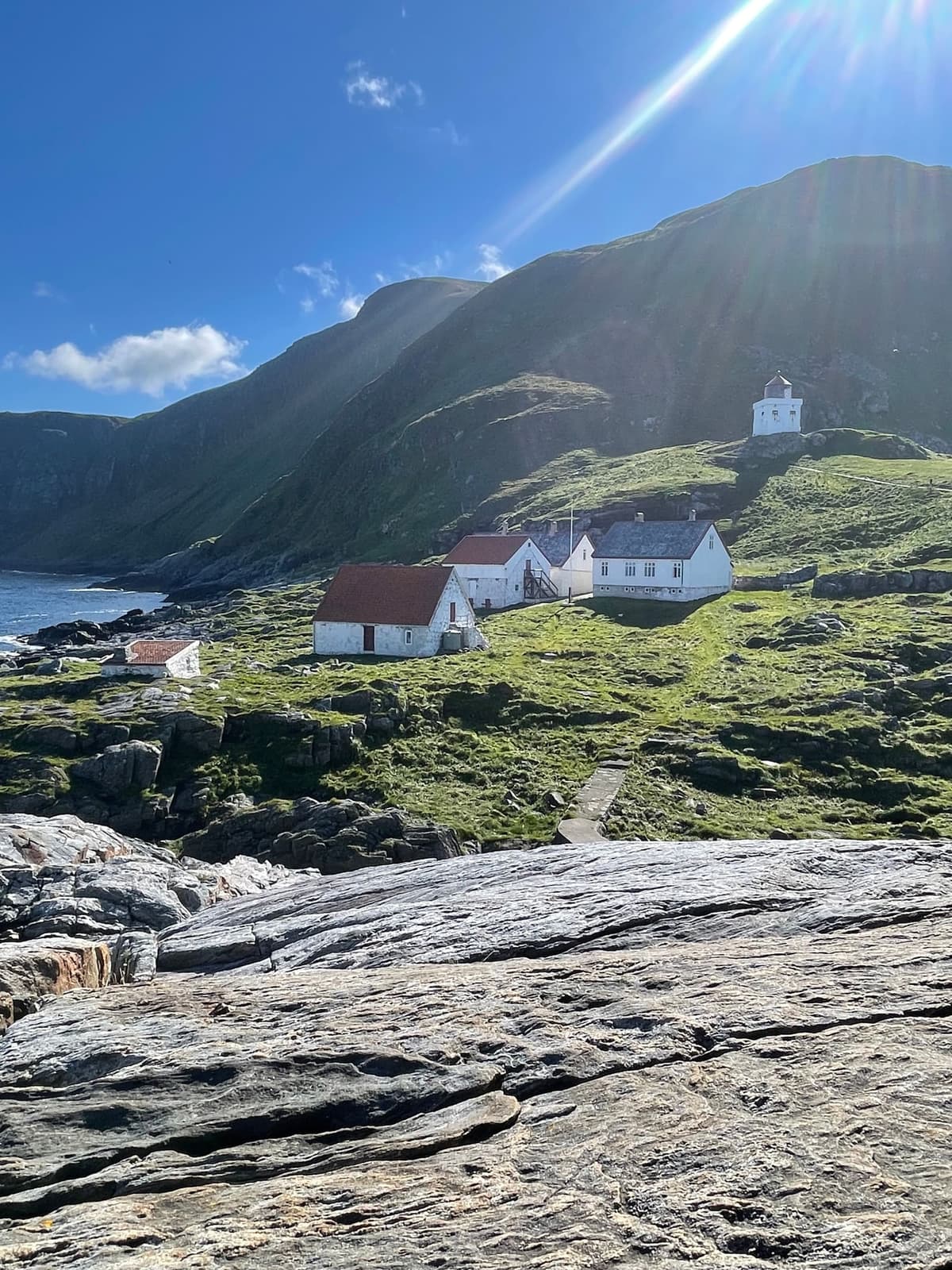 Runde fyr med fyrvokterboligene, omgitt av grønne fjellsider ytterst mot havet